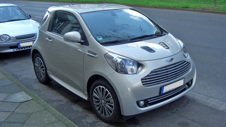 A silver Aston Martin Cygnet parked on the roadside with lush grass in the background.