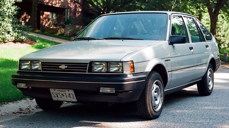 A sliver 1988 Chevrolet Nova liftback parked on the roadside with trees and a building in the background