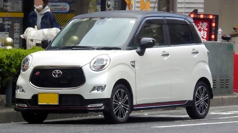 A white Toyota Pixis Joy S with a black top parked on a street in front of a building.