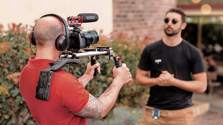 A cameraman films a presenter with a Rode VideoMic NTG Shotgun Microphone attached to his camera.