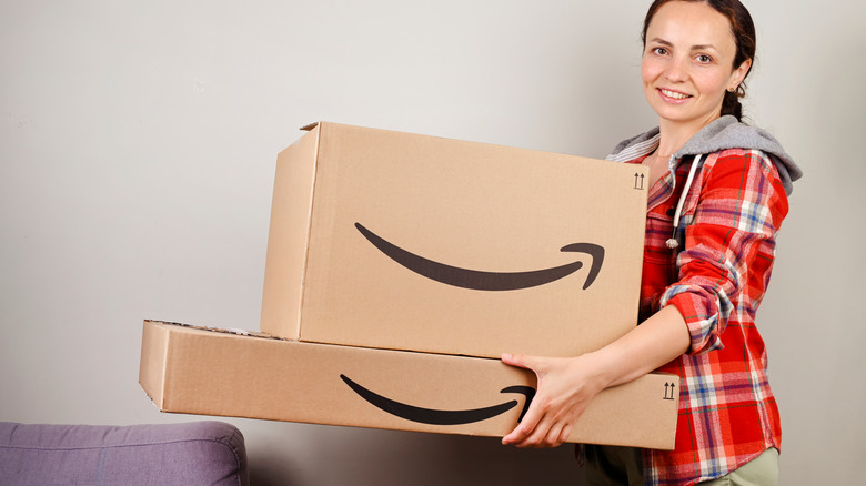 A smiling woman with brown hair in a plaid fleece holds two large Amazon boxes.