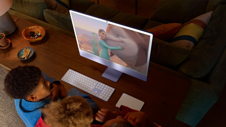 A 24-inch iMac in purple color lying on a desk in front of two children