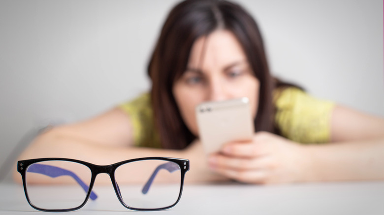 Blurry image of person staring at smartphone in the background, in-focus glasses in the foreground