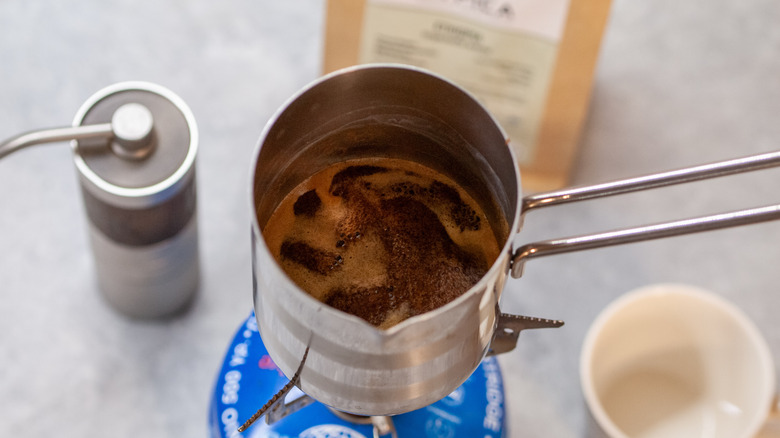 Turkish coffee brewed in Cezve over a gas stove, with a hand grinder, specialty coffee, and a cup in the background