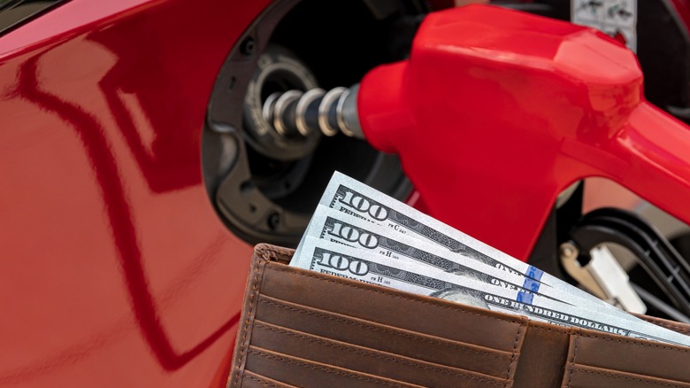 A gas pump nozzle inserted into a fuel tank fill port and a wallet showing three $100 bills