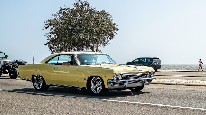 A yellow 1965 Impala Sport Coupe driving on a coastal road, front 3/4 view