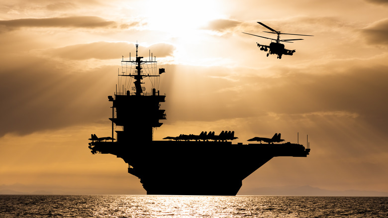 An aircraft carrier in the ocean, with a helicopter above it and airplanes on deck.