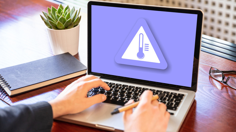 Office worker hands seen atop a laptop device with a high temperature warning sign displayed on the screen