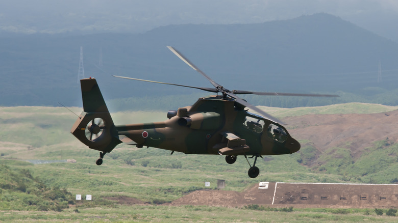 A Kawasaki OH-1 helicopter with a camouflage pattern flies above grassy hills.