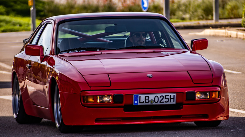 A red Porsche 944 drive by a man with a mustache and soul patch.