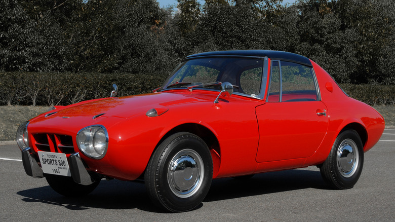 A red 1965 Toyota Sports 800 in a parking lot with leafy trees in the background.