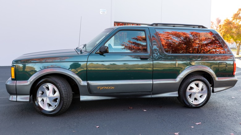 Side profile of a shiny green-on-gray 1993 GMC Typhoon, pictured against a white building, with the orange leaves of a nearby tree reflecting from the windows.