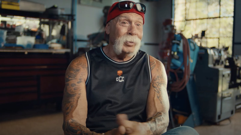 Paul Teutul Sr. in red cap and black tank top with sunglasses on forehead in workshop talking to camera