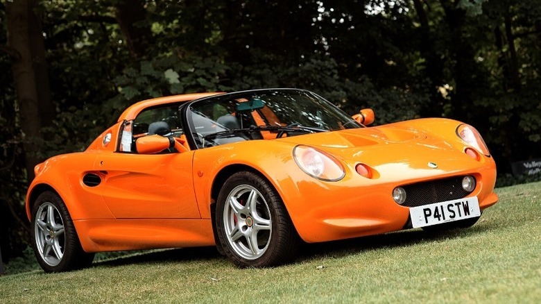 An orange Lotus Series 1 sits on grass in front of trees