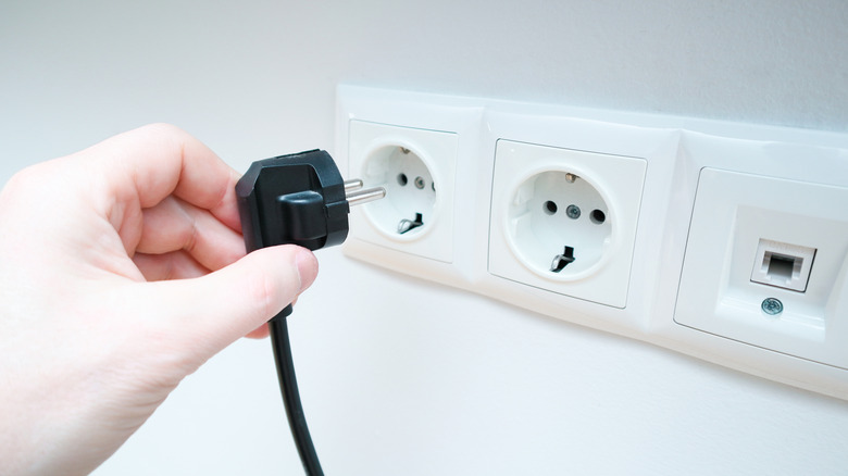 A hand holding an electrical plug next to the socket on a white wall