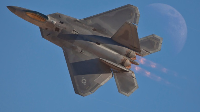 An F-22 Raptor burning its full afterburners over blue skies with the moon in the background