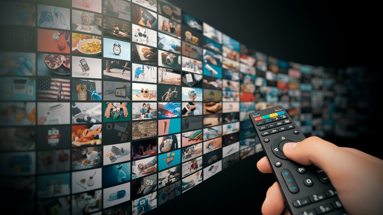 hand with TV remote pointed at a large "wall" of screens displaying various channels