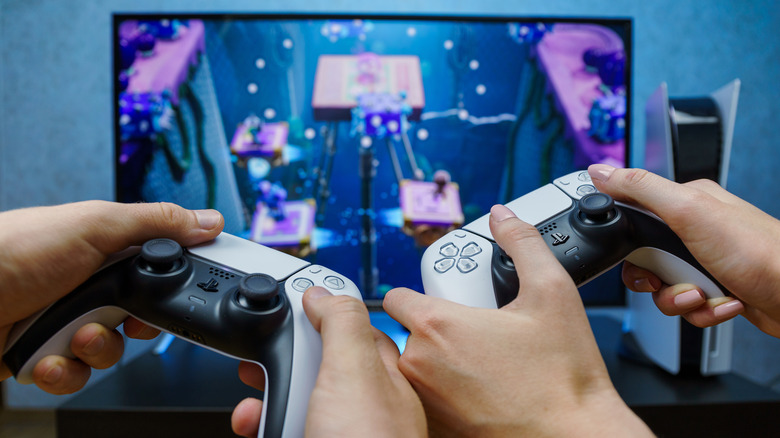 Two pairs of hands holding PS5 controllers in front of a TV