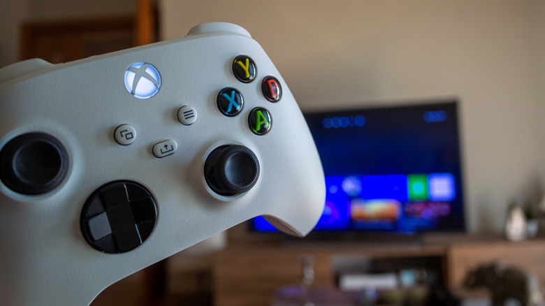 A hand holding a white Xbox controller in front of a TV.