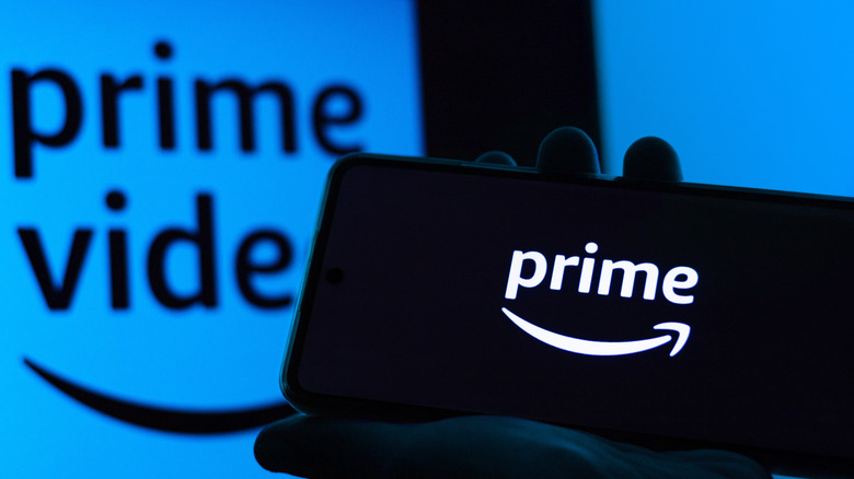 A person is holding a phone with the prime logo on it in front of a screen that has the prime video logo