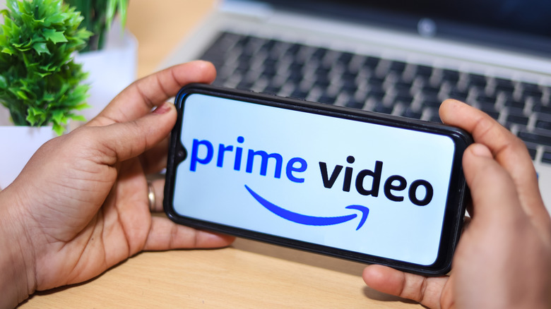 A person is holding a smartphone with two hands that bears the prime video logo in front of a laptop