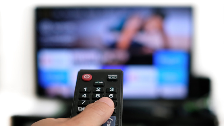 photo of a hand holding a TV remote, the a blurred TV in the background showing a streaming platform menu