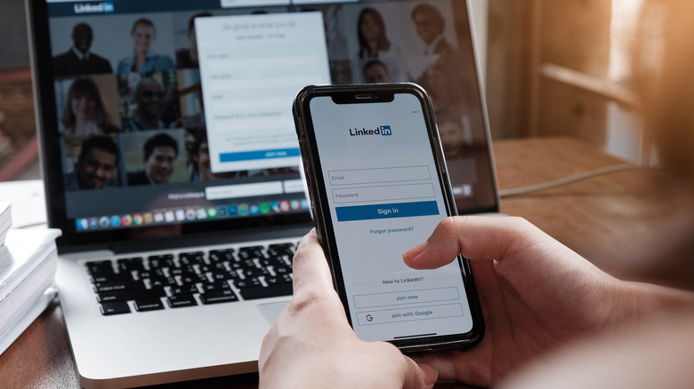 hands holding an iPhone in front of a Mac computer, with both devices displaying LinkedIn on their screens