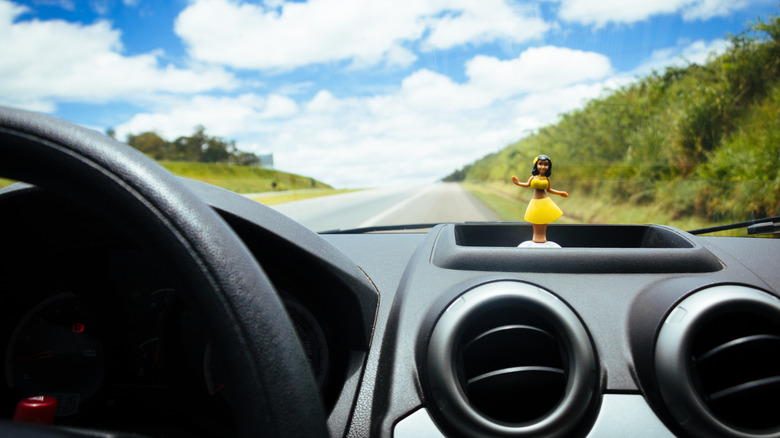 A hula girl doll sits on a car's dashboard as it drives down the road.