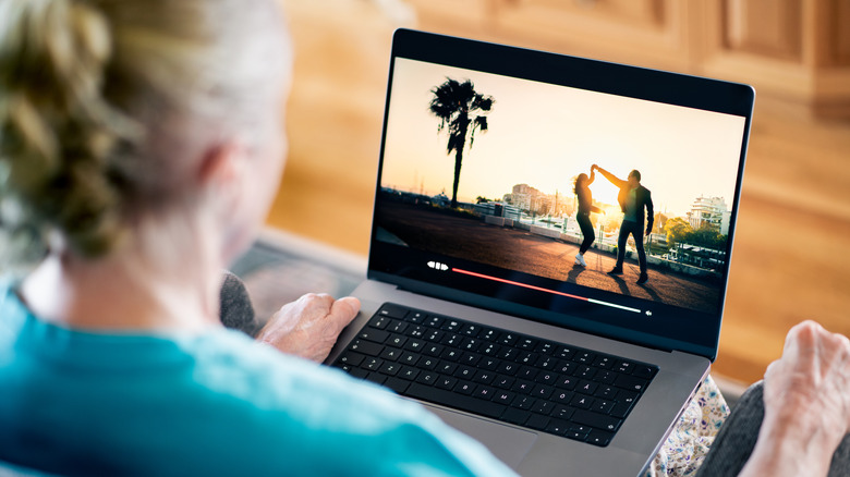 A woman seen from the back, is watching a movie on a laptop. The screen shows a couple in silhouette dancing by a beach near a palm tree.