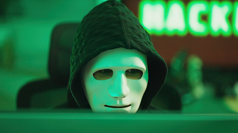 Image of a person sitting behind a computer monitor wearing black hoodie and a white mask to disguise their features. The background is blurred and green and the word HACK can be seen in green neon lettering.