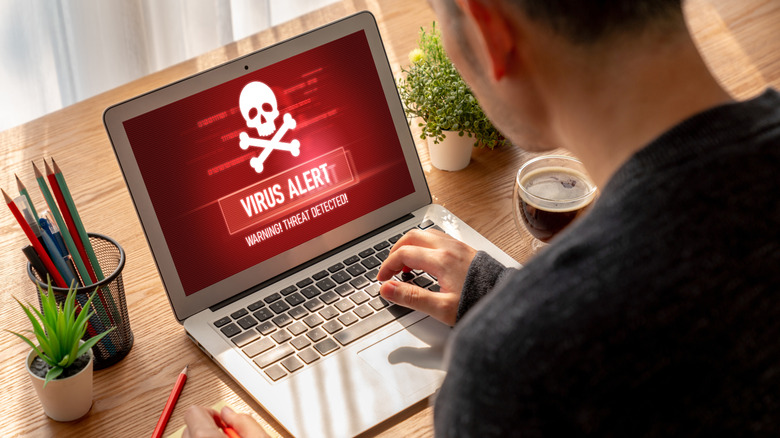 A man is sitting in front of a laptop displaying the words "Virus Alert. Warning! Threat Detection". The laptop is ona  wooden desk where there is also a cup of coffee, a pencil pot and several small pot plants.