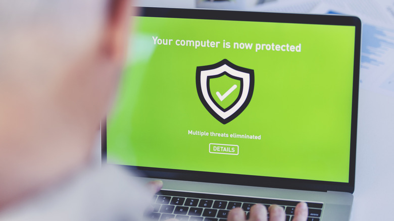 A laptop screen displays the words Your Computer Is Now Protected with a shield logo on a green background. You can see the out-of-focus back of the head of the user in the foreground.