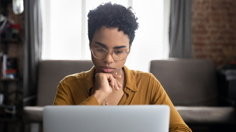 Serious woman in glasses sits at a desk staring at laptop screen. She has black hair and a brown shirt and is resting her hand under her chin.