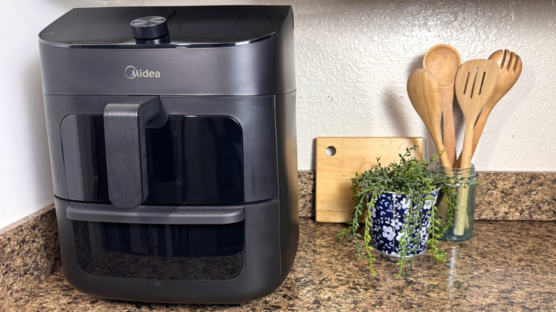 The Midea Two Zone Air Fryer on a granite countertop with wooden spoons and a green plant to its right