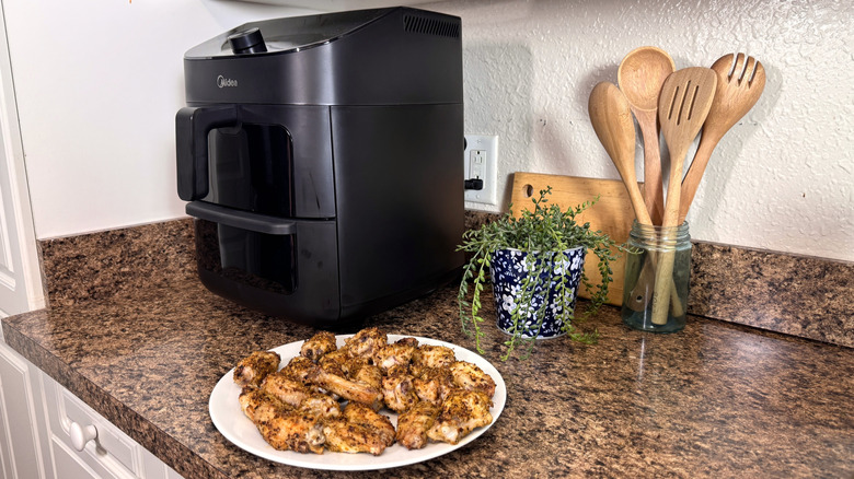 The Midea Two Zone Air Fryer on a granite counter with wooden spoons and a green plant to its right along with cooked chicken wings on a white plate.