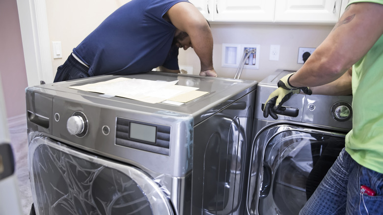 Two appliance delivery men are installing a new washer and dryer in the laundry room of home. One of the repairman is making connections in the back of the machine while his helper watches him. The new washer and dryer are energy efficient