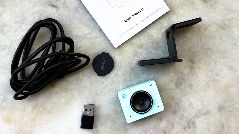 Obsbot Meet 2 webcam with USB-C cable, mount clip, lens cover, and user manual lying on the table surface.