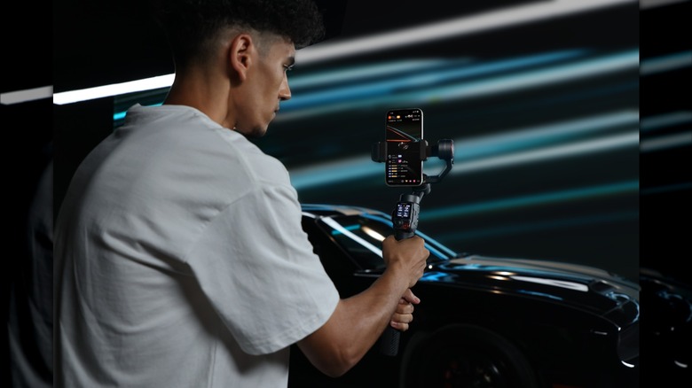 Man in white t-shirt using the Hohem iSteady M7 smartphone gimbal to film a black sports car