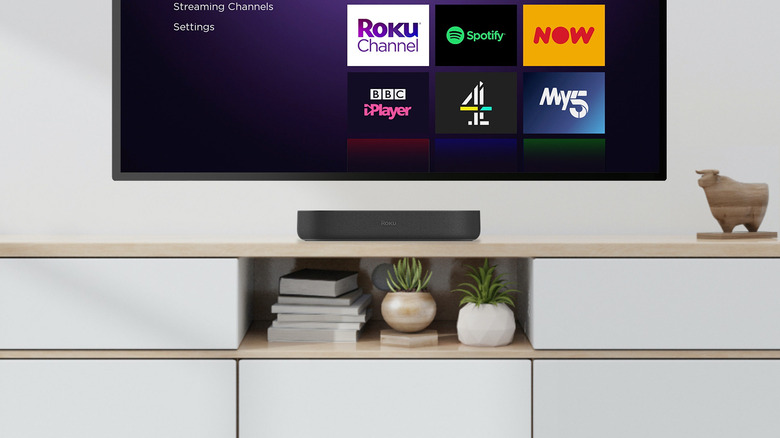 A Roku Streambar on furniture with a TV hanging on the wall