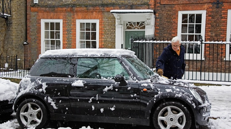 An old man removes snow from a parked Mini Cooper