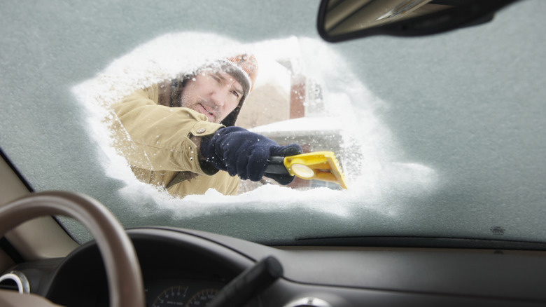 A man scrapes a voluminous amount of snow off the windshield as viewed from the inside of the car