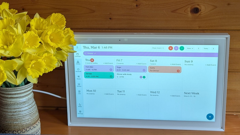 A Skylight Calendar displaying the weekly layout with several events added. It is on a shelf in front of a wooden wall next to a vase of daffodils.