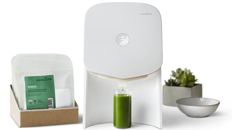 A white Juicero with a full glass of juice and juice packs to its left.