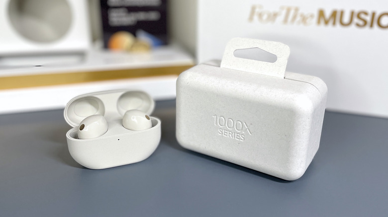 WF-1000XM5 earbuds in charging case next to their box, with "For The MUSIC" review sample packaging in background