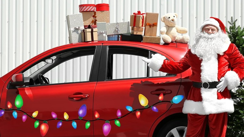 Santa stands with a car that has Christmas-light decals on the side and gift boxes and a teddy bear on its roof