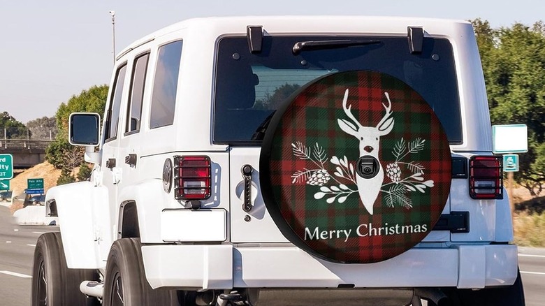 A Christmas car tire cover shows a white deer and pine cones on a red and green plaid background with the words Merry Christmas