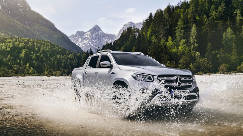 Mercedes-Benz X-Class pickup truck driving through a water puddle with a forest and mountains in the background