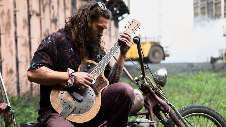 Jason Momoa playing guitar on an old motorcycle