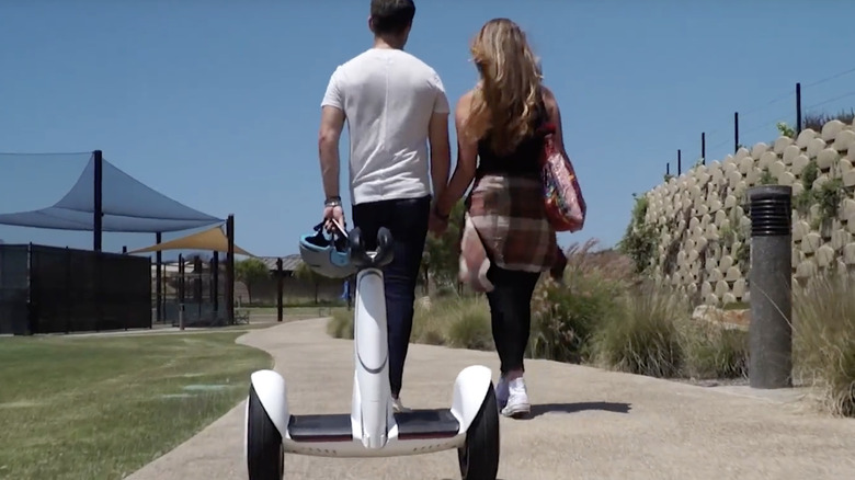 Couple holding hands walking on sidewalk ahead of Segway S-Plus scooter