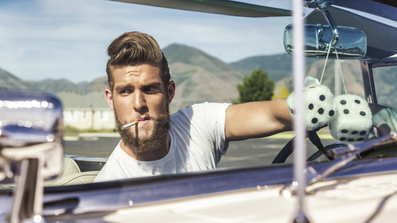 A man with a 1950s pompadour hair driving an open top convertible car looking with fuzzy dice looking to race.